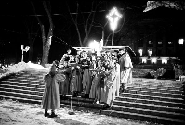 Night view of Truax Chapel Choir members standing on the steps at the State Street entrance of the Capitol Building singing Christmas carols to passers-by while they shop on the Capitol Square.