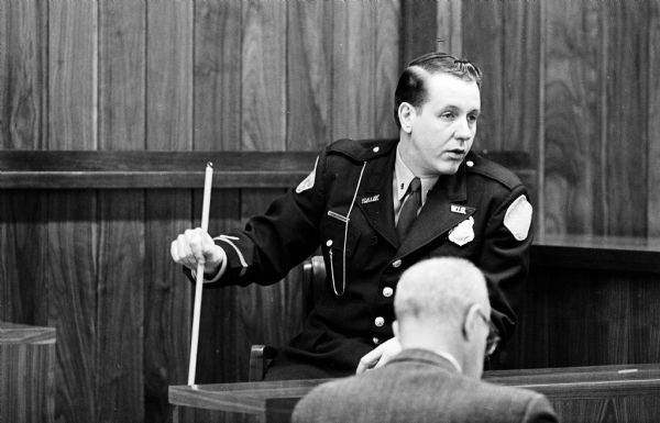 Sauk County Policeman Fred Bayer, in uniform, using a pointer stick while testifying at the trial of William Welter, Lawrence Nutley, and Richard Nickl. They were accused of slaying Patrolman James Lantz, Baraboo, and wounding Lake Delton Police Chief Eugene Lee Kohl in an early morning gun battle in front of the Lake Delton village hall.