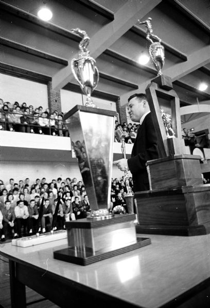 Dave Brown, coach of the Edgewood High Boys Basketball team, attends a victory rally after winning the State Catholic Basketball Championship. In the foreground are two large trophies.