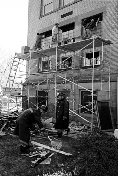 Employees of the Monona Masonry Company continue to install glass block windows at the Madison Board of Education building, 351 W. Wilson Street, while firemen battle a blaze that was started by a workman's molding torch.