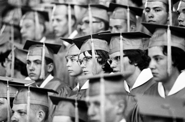 The image centers on five unnamed faces in the crowd at the 1962 East High School graduation ceremony. Everyone is wearing graduation robes and mortar board hats with tassels.
