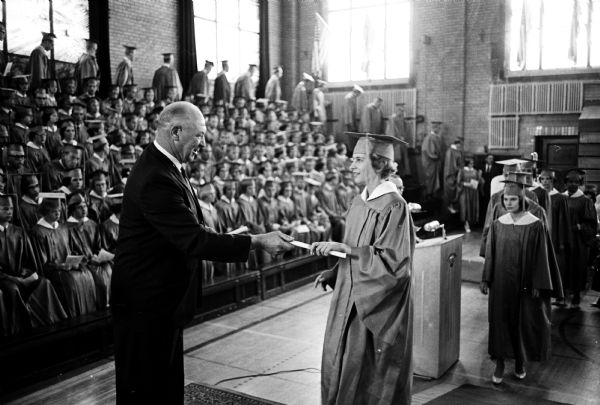 An unnamed graduate receiving a high school diploma as the audience is looking on at the 1962 East High School graduation ceremony. Presenting the diplomas is school board member Arthur (Dynie) Mansfield. Mansfield was also a U.W. education professor and baseball coach.