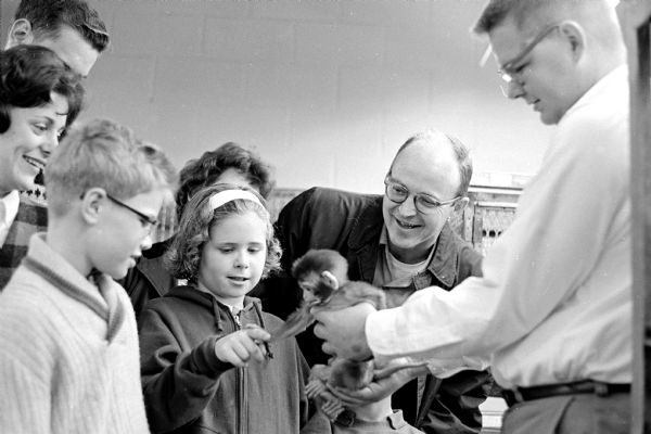 An associate of U.W.'s "World-Famous" Primate Laboratory holding out a two-month old monkey for a young smiling girl to pet as her parents are looking on.