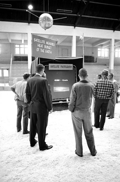 A small group of men viewing a satellite photo exhibit displayed at the U.W. Stock Pavilion during the Open House. Suspended from the ceiling above them is a life-sized model of a satellite.
