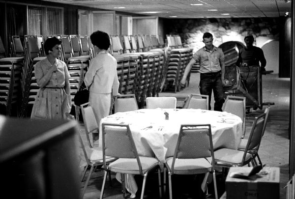 Interior view of a set dining table in front of workmen and stacked chairs at the new Park Motor Inn on Madison's Capitol Square during the rush to open for business.