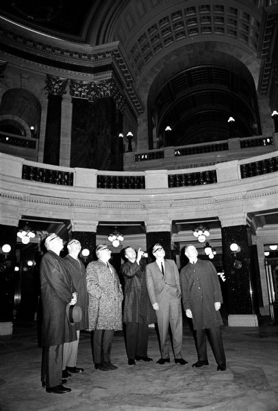 Six men are shown standing in the Wisconsin State Capitol Rotunda. All men are looking upward as one man points to the top of the Rotunda.
