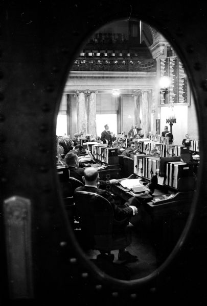 Assemblyman Paul R. Alfonsi (R-Minocqua) is framed by the oval window in a door of the Assembly chamber as the majority party floor leader addresses the Assembly regarding a bill to repeal a three-month old law requiring taxpayers to pay a $2 filing fee when filing state income tax returns.