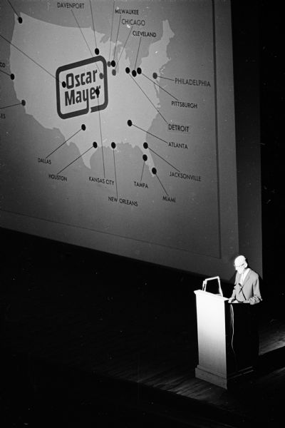 Oscar G. Mayer Jr., president of the Oscar Mayer Company, at a podium on the stage of the Orpheum Theatre. On the screen is a large map of United States with a logo of Oscar Mayer. He is giving a report of the company including a premier showing of the film "Bring Home the Bacon" to employees of the 80 year old Madison plant.