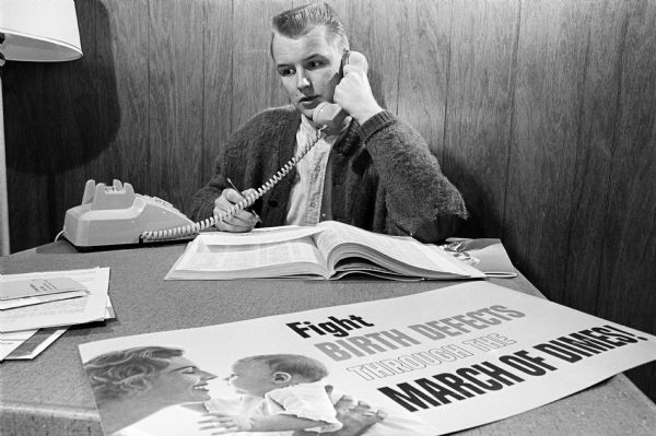 Dick Collman, soliciting contributions by telephone, is one of the "Polio Pioneers" celebrating the 10th anniversary of the Salk vaccine trial by helping with the March of Dimes campaign. The slogan as seen in the poster on his desk is "Fight Birth Defedts through the March of Dimes" and represents one of the many areas in which the March of Dimes supports research.