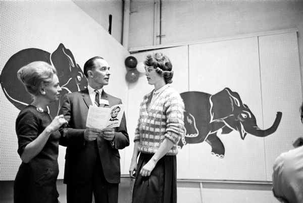 Republican candidate for the U.S. Senate Wilbur N. Renk chatting with party volunteers Mrs. David Orfan and Kay Smith at the annual Lincoln Day dinner. Renk is holding an open pamphlet. Behind them is a poster of a running elephant.
