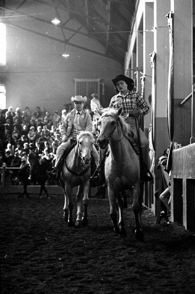 A young woman astride a horse during the Little International Livestock and Horse Show at the U.W. stock pavilion. In the background are a crowd of people and another horse and rider.