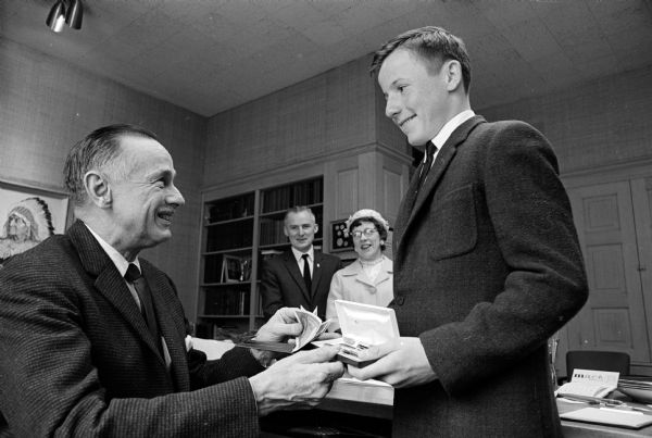 Kirby Toberman, The Wisconsin State Journal's 1964 Young Columbus, is shown receiving a passport holder and gift pen and pencil set from State Journal Publisher Don Anderson. Kirby, a 13-year-old eighth-grader from Cassville, won a subscription contest for the paper. In the background are his parents, Mr. & Mrs. David Toberman, who came to see him off for Italy that morning.