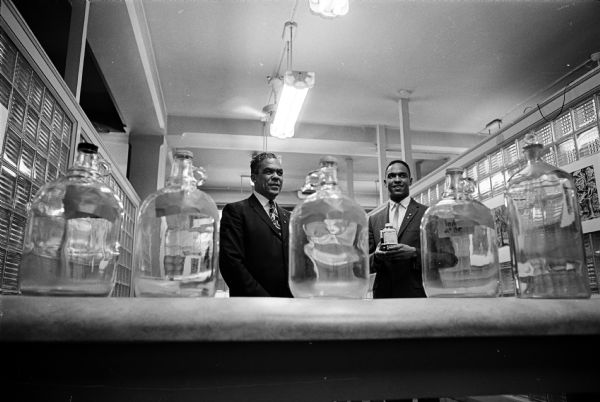 Harry L. Hamilton and his son Harry Jr. pose with five large glass jars that symbolize the five gallons of blood they have donated to the Dane County Red Cross. They are among the many two-generation families that donate on a regular basis. Harry was managing editor of the <i>Agronomy Journal</i>, and was married to Velma Bell Hamilton. Both Harry and Velma were active in civil rights issues and in aging issues in Madison.