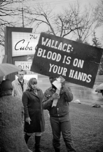 Three people of the 18 protestors outside of the Cuba Club restaurant. A large protest sign states, "Wallace:  Blood is on your hands." The protestors were members of the Madison chapter of the Congress on Racial Equality (CORE). Inside, the Madison Exchange club's invited speaker, Alabama governor George Wallace to give a speech at the Cuba Club restaurant for 180 of their members. Wallace and his contention that the pending civil rights bill will "destroy the constitutional rights of everybody" were met with applause.