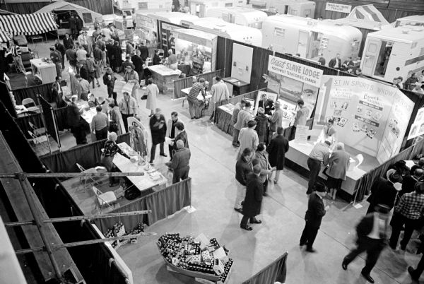 An overhead view of the Madison Sports-Boat-Vacation show's opening night at the fairground. In the foreground a crowd of people are viewing booths with exhibitions. Tent and house trailers are seen in the background.