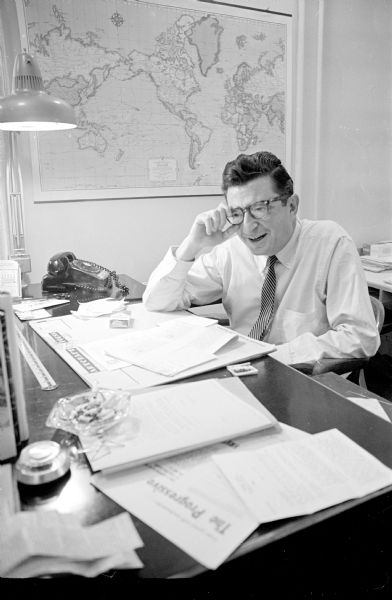 An informal portrait of Morris Rubin sitting at his desk, decked with a glass ashtray, pack of cigarette and matchbook, rotary-dial telephone and papers. Since 1940, Rubin has been the editor of <i>The Progressive</i>, a liberal magazine published in Madison. The magazine was founded 55 years earlier (1909) by Robert La Follette, Sr.
The year this photograph was taken, the publication had subscribers in 116 countries, and was exerting international influence.