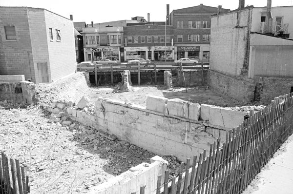Image shows an empty lot on the 400 block of State Street across from Ella's Deli (425 State Street). In the lot are remnants of basement walls and brick rubble. Across the street is the Catholic Info Center, 427 State Street; Ella's Deli, 425 State Street; Bluteau's Wholesale Meats, 421 State; and Martin's Taylor Shop, 419 State Street. The lot is currently Lisa Link Peace Park. Five cars can be seen parked on the street.
