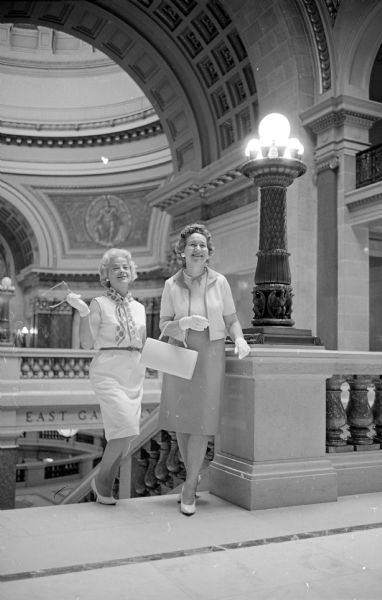 Two women wearing dresses and white gloves are posing by a railing inside the Wisconsin State Capitol building. They are wives of military reserve officers and members of Reserve Officers Assn.-Ladies (ROAL).  Their names are Mildred, wife of Maj. Walter Magill; and Mrs. Alfred Plaenert, wife of Col. Plaenert.