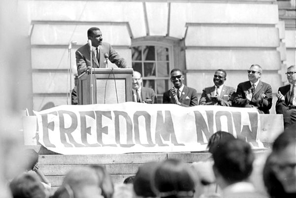 Rev. Fred L. Shuttlesworth, president of the Alabama Movement for Human Rights and a close associate of Rev. Martin Luther King, speaks to a rally of 400 people, held on the State Capital steps, calling for the passage of the Federal Civil Rights Bill. A "Freedom Now" banner is shown in front of the podium.