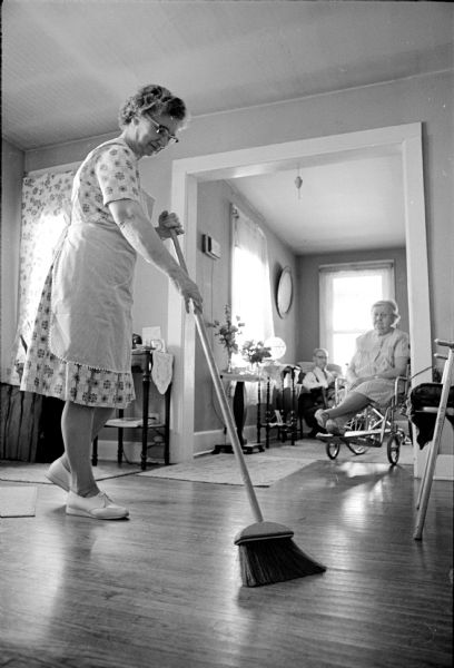 Marie Ackley is sweeping the floor of the apartment of Mr. and Mrs. Alfred Lund. She is working as a health aide in a new Visiting Nurse Service program and works in eleven different homes each week.