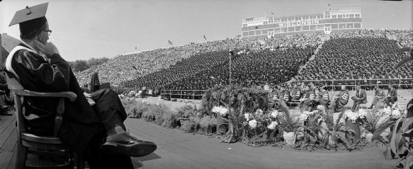 University of Wisconsin's 111th Commencement in Camp Randal stadium.  View is a panorama looking from the stage at the Class of 1964 graduates and their guests seated in the stands. In the foreground is a seated man in a cap and gown looking at the crowd. Between them are many flower bouquets.