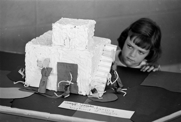 Marianne Davies, a third grader, looks over a work created by a group of fourth graders which is on display at the First Baptist Church Children's Day art exhibit on display in the church library. The work represents a home like the one Jesus lived in as a boy and was built out of a small corrugated box covered with a paste mixture of flour, salt, and water and based on pictures they had seen of family dwellings in the time of Christ. The figures are dressed according to the style of the era.