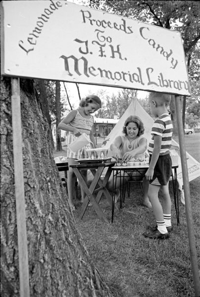 The children of Donald and Jeanne Jacobs selling lemonade, candy, gum, and other goodies to aid the John F. Kennedy Memorial Library fund. Karen, 9, is pouring lemonade while Patty, 13, is straightening the display table. Dave, 4, is supervising.
