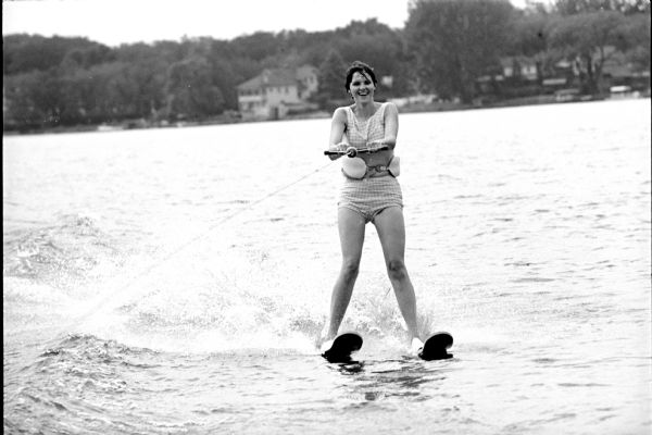 View from tow boat towards a woman, named Anne, water skiing. In the background are houses along a wooded shoreline.