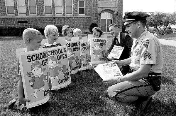 Arthur Wichern, public relations director for the Wisconsin division of the American Automobile Association (AAA), and Sergeant Jack R. Geiwitz, Monona police officer, inspect safety posters worn by young "motorist reminders." The young sandwich-people are, left to right, James and Katherine Johnson, children of Mr. and Mrs. Bruce N. Johnson, 5506 Pheasant Hill Road; Valeria Hoffmann, daughter of Mr. and Mrs. Wayne C. Hoffmann, 203 Stone Terrace; and William and Cynthia Schwartz, children of Mr. and Mrs. Myron C. Schwartz, 5304 Pheasant Hill Road. Taken in front of the Nichols School on Monona Drive.