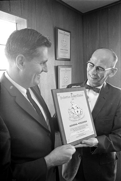 Two men are holding a special award which reads: Pedestrian Safety Citation 1963. Monona, Wisconsin is cited for its record of No Pedestrian Deaths as reported to the AAA Pedestrian Program Appraisal while meeting AAA standards of program performance. 3 Year Achievement. Presented by the American Automobile Association. The men are not identified.  