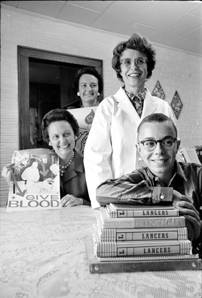 Mark Newman is shown sitting behind a stack of books as he returns to La Follette High School three weeks after radical surgery for a brain tumor. Behind him in a white surgical coat is his mother, Rosemary Newman, a Red Cross volunteer nurse. At left is Ellen Rosten (seated), and Margaret Kilgust Co-Chairmen, of the Monona Grove Red Cross Volunteers.