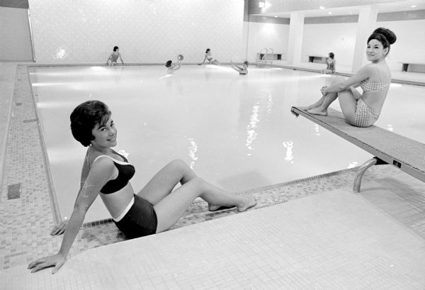 One of a series of photographs showing the luxurious features of private dormitories on campus, two residents of Lowell Hall, 610 Langdon Street, are shown lounging at the dorm's swimming pool wearing bathing suits. Left is Pat Factor, Glencoe, Illinois. Perched on the diving board is Vicki Morris, Davenport, Iowa.