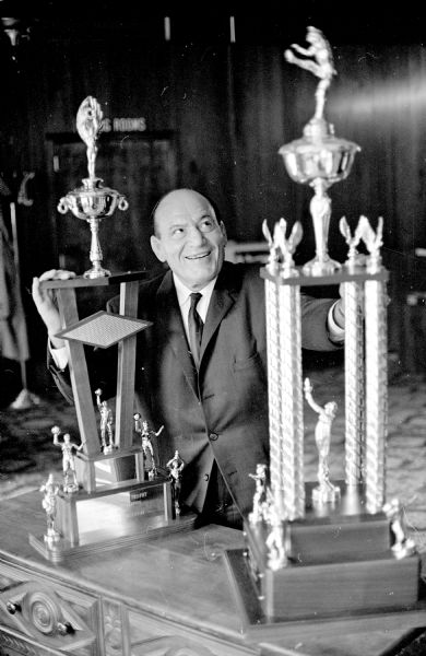 Madison wrestling promoter Jimmy Demetral posing proudly between two large ornate trophies that he would provide and present annually to the player voted most valuable on the Badger football squad, and to the free throw champion of the Badger basketball team.