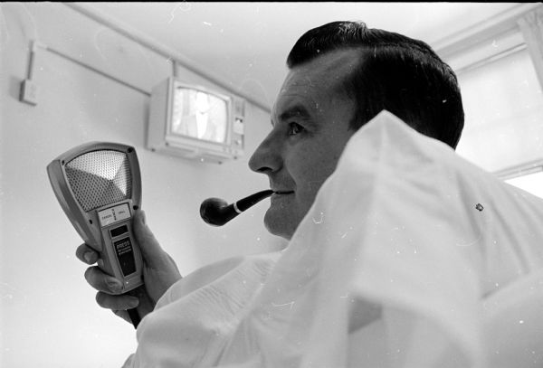 View of Joe E. Miller with his head on a pillow trying out a new hand held "communication system unit, which allows him to  change the channel on the television or  radio, adjust the volume, or turn it off. The middle button allows him to call and communicate with his nurse." In the background is a wall mounted television. A tobacco pipe is in Miller's mouth. The new system was installed at Methodist Hospital.