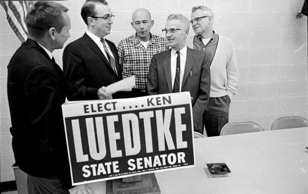 Two politicians and three labor officials are standing behind a campaign sign that says: "Elect Ken Luedtke State Senator." Exchanging greetings are William McCullough, First ward alderman and chairman of the reception; Luedtke; Claire Soule, president of Laborers Local 464; William Eley, vice-president of Painters Local 803; and Jack Fitzpatrick.