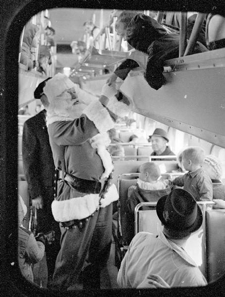 Santa Claus is shown in the aisle of the Santa Express Train shaking many hands extended in this double decker train car, one of eight cars from the Milwaukee Road passenger train. 6,000 children rode from Madison to Stoughton and back again.
