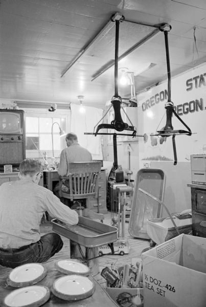 View of two men, with their backs to the camera, are sitting in a workshop at Oregon State Farm painting toys. Pieces of freshly painted, disassembled toy wagons are around the room. The news story says the donations are intended "for the children at Lac Court Orielles Chippewa Indian reservation near Hayward." Also several truckloads of toys were collected by the congregations of six Oregon (Wis) area churches.
