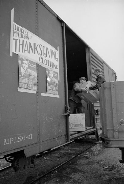 Two men are transfering boxes of donated clothing from a truck into the open side of a railway freight car parked at Cross Plains. The two men are Frank Hoffer, and the Rev. Father Joseph Braig, both of Waterloo. Taped to the side of the boxcar is a large poster that reads: "Diocese of Madison Thanksgiving Clothing." Below that are two smaller identical posters with the picture of a young boy and the words: "Thanksgiving Clothing Collection." The effort was sponsored jointly by the Catholic Relief Services and the National Catholic Rural Life Conference.   
