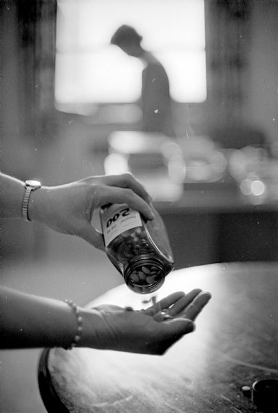 In the foreground, a woman's arms, with a bracelet and wristwatch. In one hand she is holding a large bottle of pills, and is pouring the pills into the other hand. In the background is the blurred silhouette of another woman. Taken at Mendota State hospital. The article is about residents being treated with tranquilizers, anti-depressants, and several months residency in pleasant surroundings.