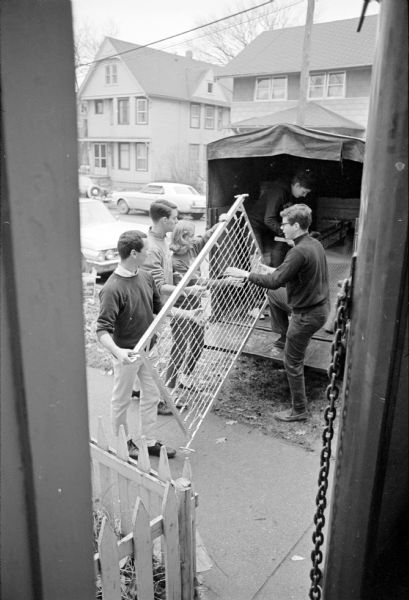 Four U.W. Greeks unloading a bed frame from a truck. The Department of Public Welfare Group Home (for women) moved from 404 W. Washington Avenue to 305 N. Livingston Street, and was assisted by volunteers from the Delta Zeta sorority and the Alpha Chi Omega fraternity. In the background and across the street are the 302, 304, and 308 N. Livingston Street addresses.