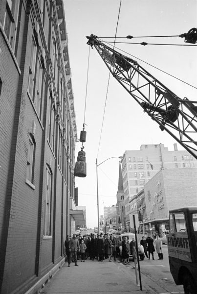 View of a crane about to swing a wrecking ball into a building wall at the 30 W. Mifflin Street and Carroll Street corner of the Square as a crowd is looking on. Behind them is the west side of Carroll Street, with four commercial buildings and a church. The caption reads: "The new 10-story building, '30 on the Square,' will be the first in Madison since 1936 to be devoted completely to offices and business suites."