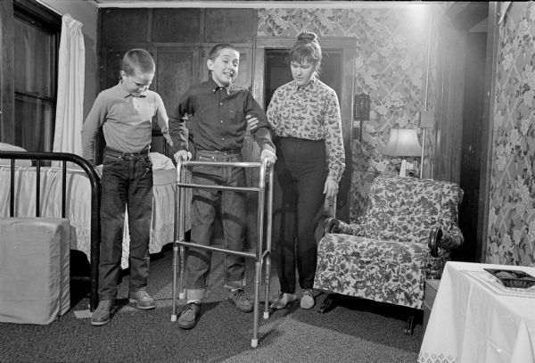 William Grab, age 12, learning to walk with the help of his mother, a boy, and a walker seven months after being crushed when a two-ton culvert rolled over him while playing in the yard of Wisconsin Culvert Company on Ingersoll Street. A playmate with him was killed.