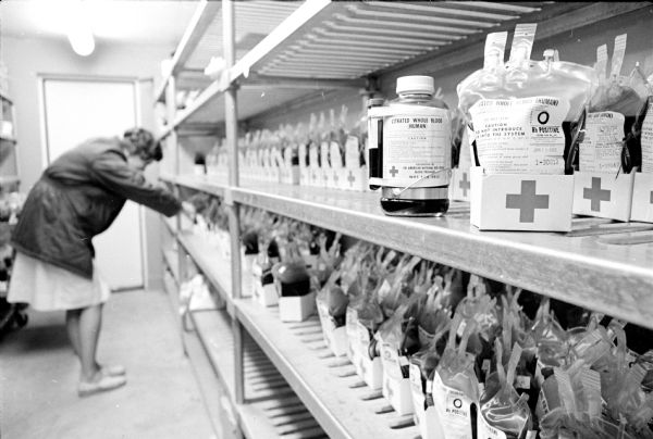 Technician Gail Stelzer checking shelves filled with new plastic blood transfusion bags at the Madison Chapter of Badger Red Cross, which recently converted from using glass blood transfusion bottles. A sample of the glass container is on the right on the top shelf.