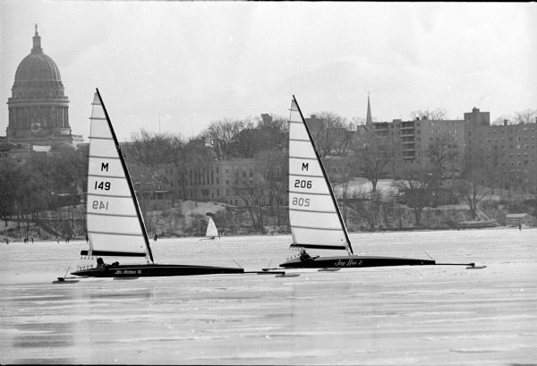 Two class A iceboats, sailed by Jack Ripp and Bob Brockel, racing on Lake Mendota. They were participating in the Wisconsin Stern Steering Association regatta. The Wisconsin State Capitol is in the background on the left. The iceboat on the left is the "Miss Fotune IV", and the iceboat on the right is the "Jay Bee 2".