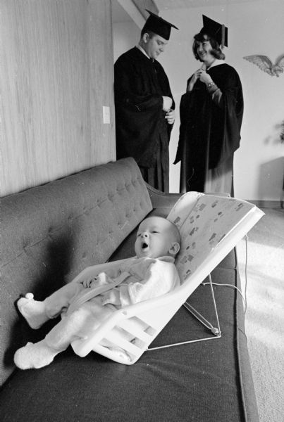 Greg and Patty Herrling are shown standing in their graduation attire on their mid-year graduation day, while their baby, Jimmy, is yawning while sitting in a infant seat set on a couch in the foreground.