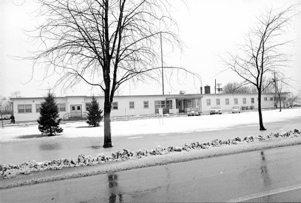 View across road towards a one-story building with three cars parked in front near the corner of E. Washington Avenue and N. Stoughton Road. The building was the Truax Field Medical Clinic and was part of the U.S. Air Force's Truax Air Base, which was vacated in June 1968.