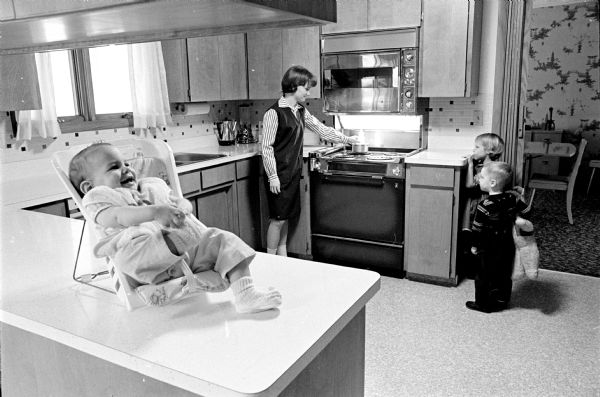 Trained baby sitter Colleen Russell, 14, heating a bottle in the kitchen of Robert and JoAnn Kessenich. In the foreground is 8 month old Nancy.  At right, observing, are Jeffrey, 2 1/2, and Cathy, 5.