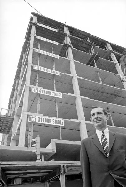 Stanly Sirotin is posing in front of the corner of the "30 On the Square" sky scraper. The low, steep angle of the photographer's camera, looking up, reveals nine stories of the building's still exposed frame. Signs on six of the floors state the floor number and its estimated completion date. Stanley Sirotin, is managing rental and construction. The new building on the Capitol Square was the first since 1936 dedicated solely to professional offices. "30 On the Square" is a 10-story glass-and marble-faced building at 10 W. Mifflin street. A Rennebohm drug store was on the ground floor.  He also developed the Pyare Square building on University Ave. in 1966.