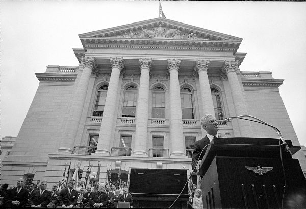 Low angle view looking up at the full height of the face of one wing of the Wisconsin State Capitol building. At lower right, behind a podium, is Gov. Warren Knowles. At lower left are politicians sitting in front of flags. The capitol was never dedicated after completion in 1917. Forty-eight years later, on July 7, 1965, it was formally dedicated, after it's exterior was chemically cleaned.