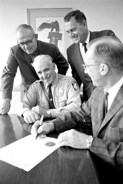Maple Bluff police marshal A.J. Taff, center, is shown sitting at a table as Madison's Mayor Otto Festge, right, is signing a document making him honorary marshal of the city of Madison. This occasion was held in recognition of his record of cooperation with the city, as well as honoring him as he retires from service. Looking on were Dane County Sheriff Vernon Leslie, left, and Madison Police Chief Wilbur Emery.